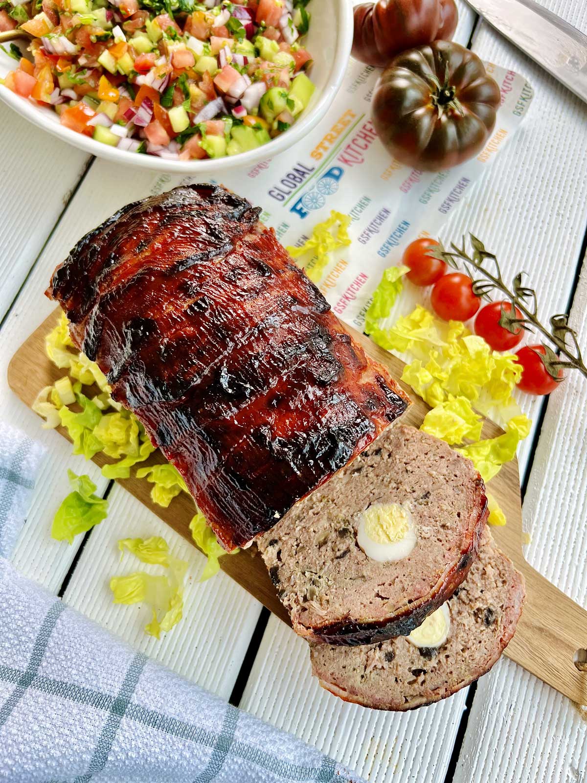 Birdsview off smoked meatloaf on a chopping board with salad, scattered around tomatoes and a bowl of salad.