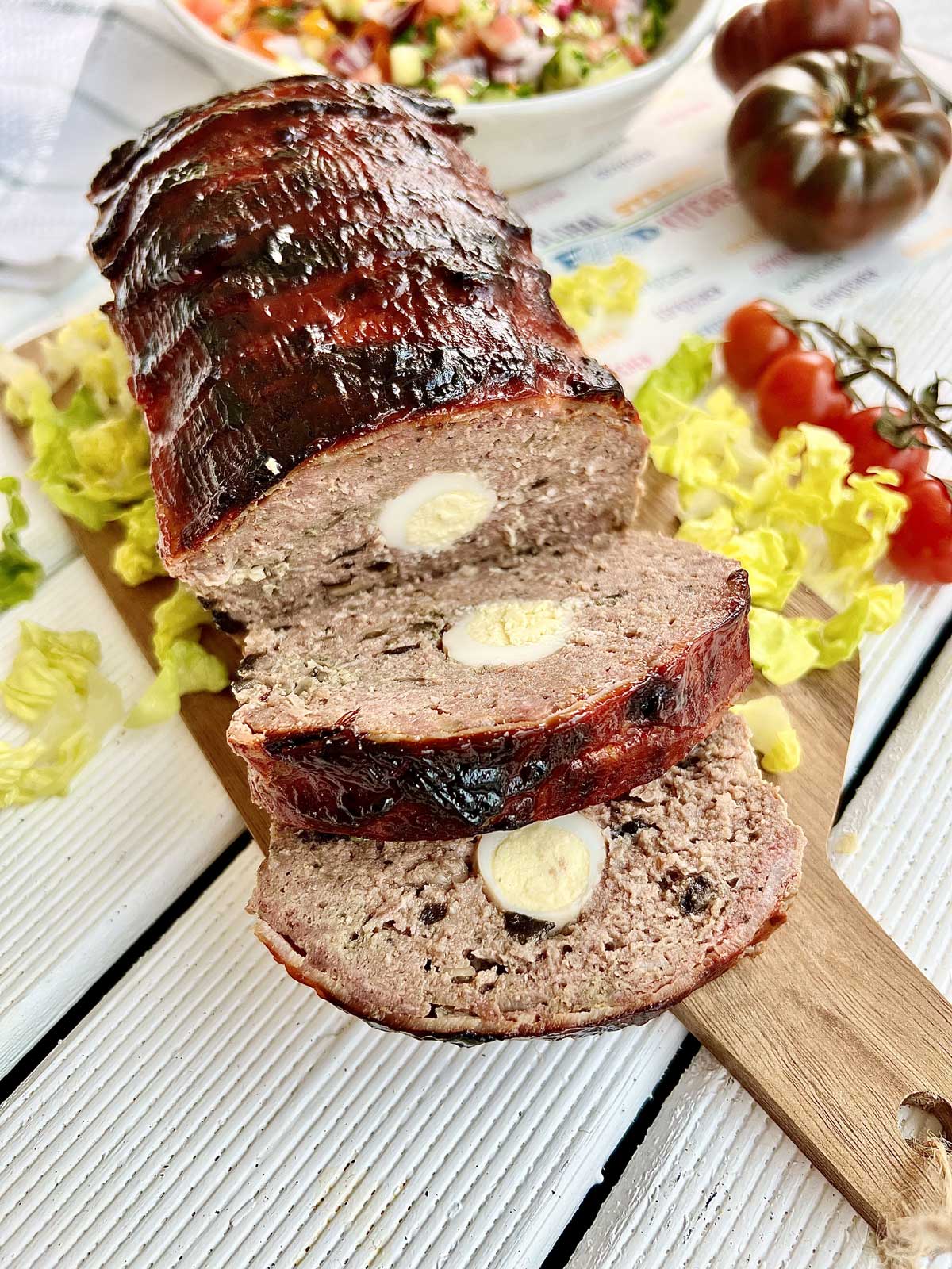 Smoked meatloaf on a wooden chopping board with salad and tomatoes around it.