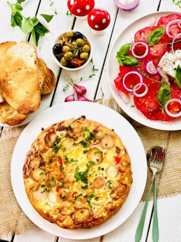 Spanish tortilla on a white plate, tomato salad on a white plate on the right mixed olives on top and toasted bread slices on smaller plate on the left