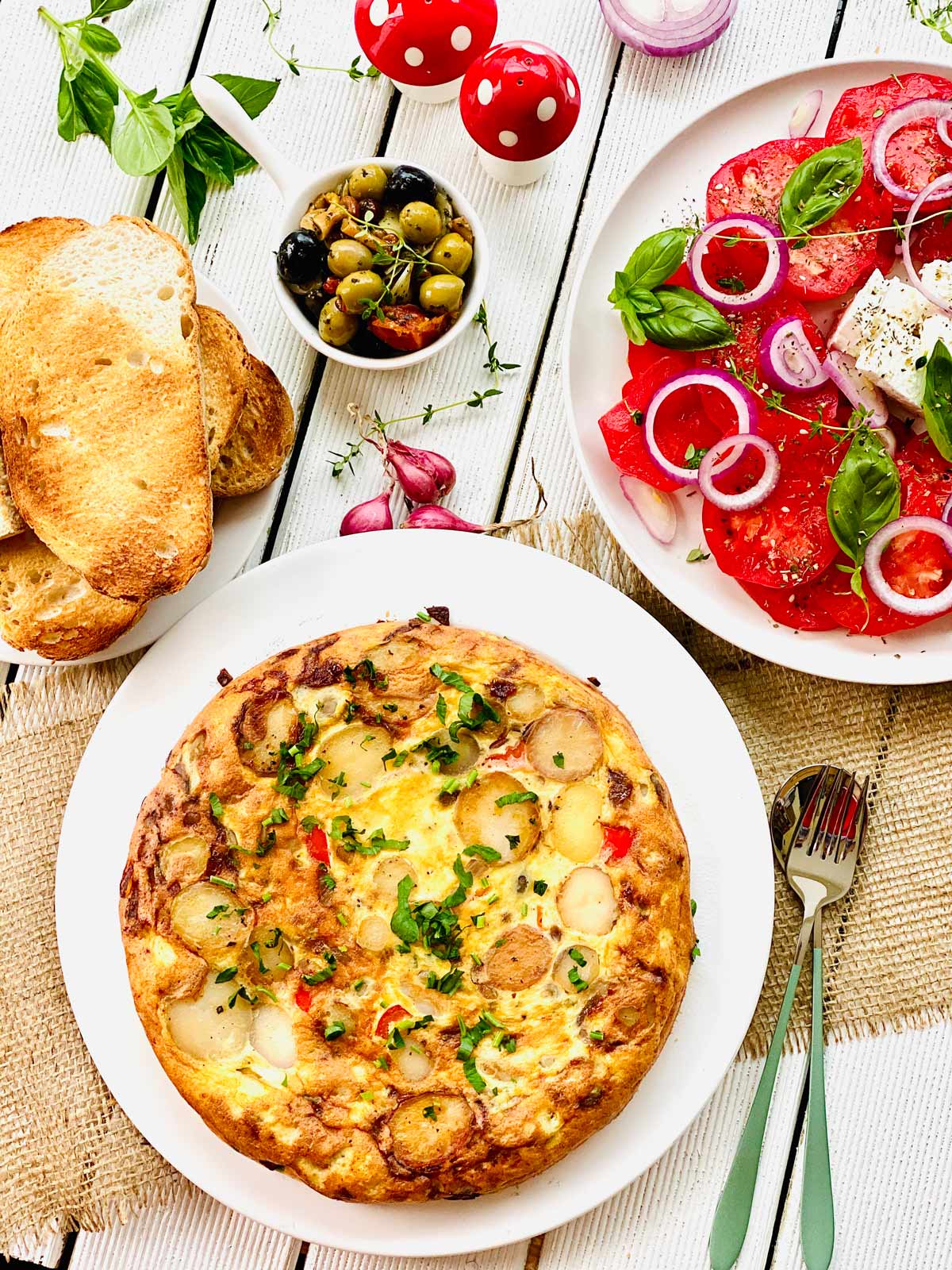 Spanish tortilla on a white plate, tomato salad on a white plate on the right mixed olives on top and toasted bread slices on smaller plate on the left.