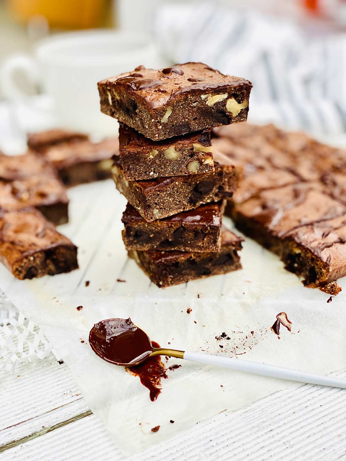 A stack of 5 protein brownies on parchment paper with the rest of the brownies scattered around and a tablespoon dipped in chocolate at the front