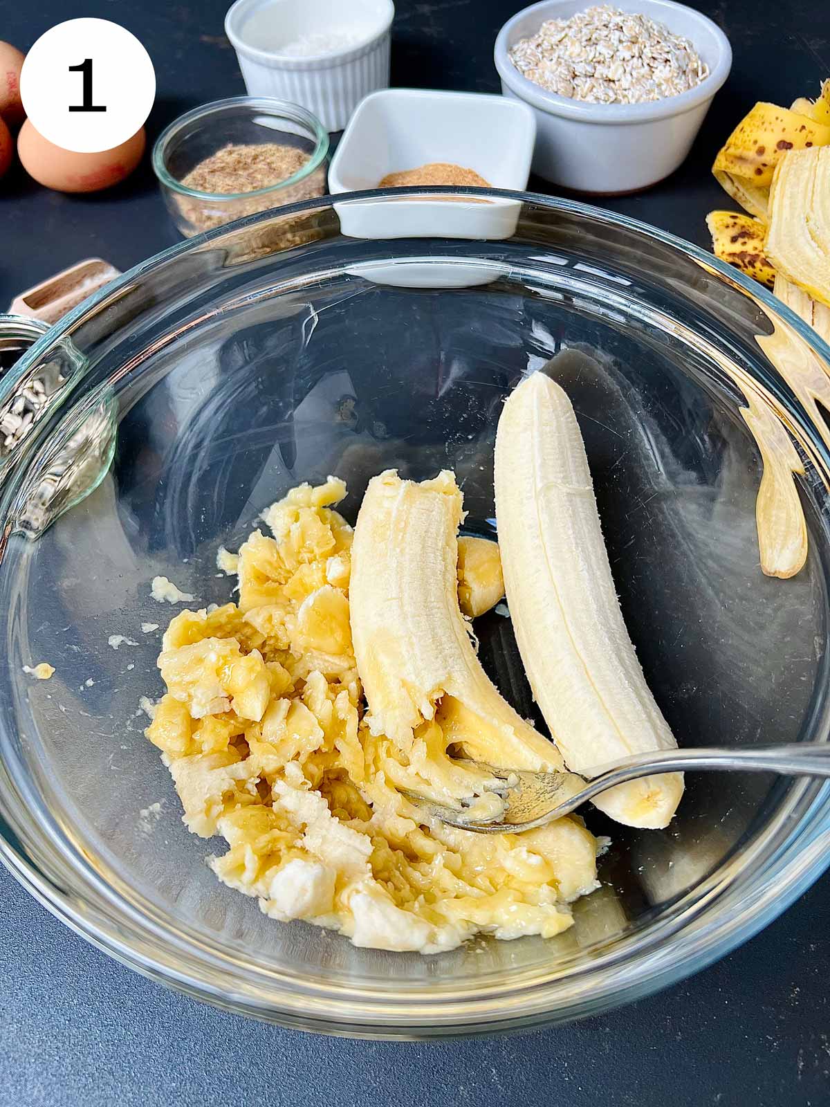 Three bananas being smashed in a glass bowl with a metallic fork.