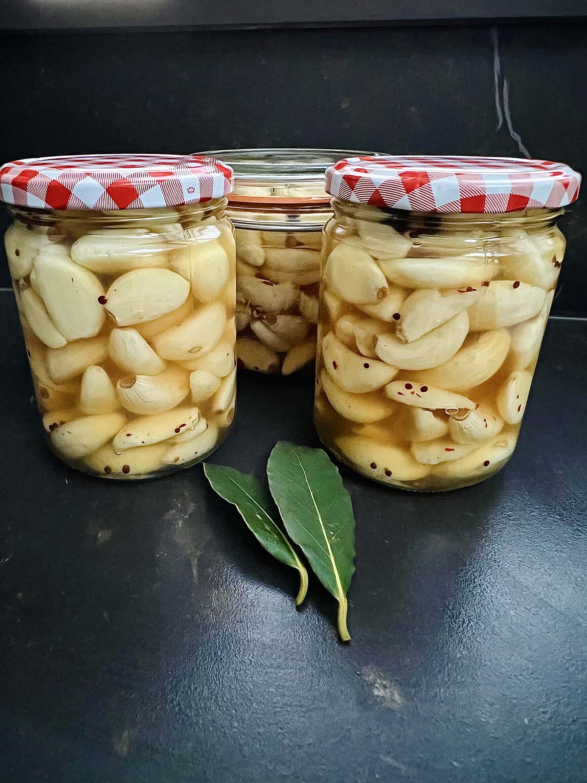 Three jars containing pickled garlic on a dark worktop and 2 bay leaves.