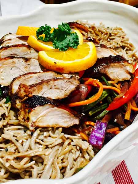 A white plate of delicious duck in buckwheat noodles, topped up with lemon slices and kale leaves.